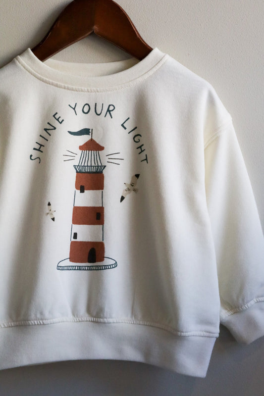 White sweatshirt with a lighthouse design and 'Shine Your Light' text on a wooden hanger.